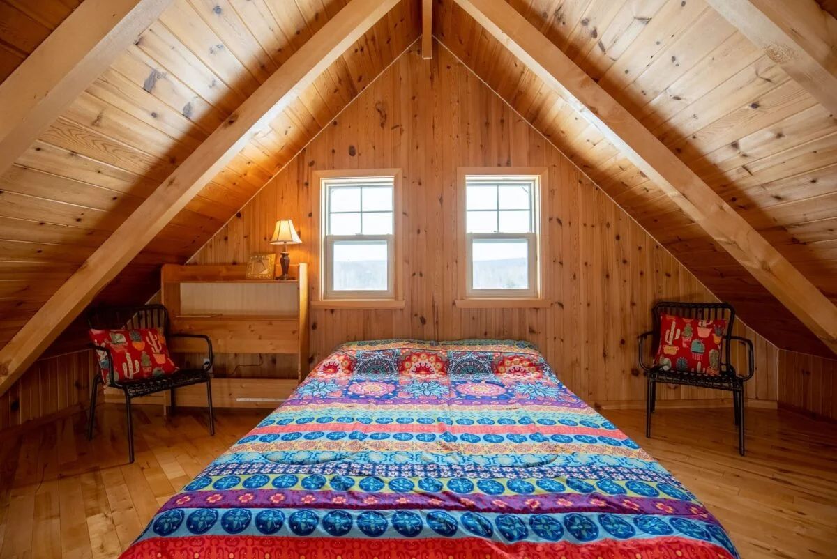 Bedroom in the attic with a double bed, wooden walls, and two bright windows.
