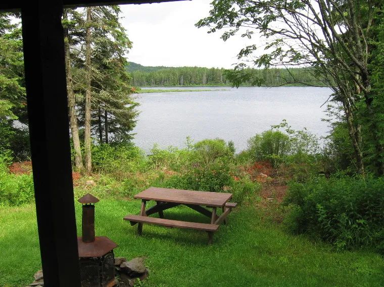 Vue sur le lac depuis la galerie du chalet incluant une table de pique-nique et un foyer extérieur.