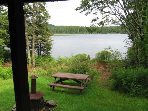 Vue sur le lac depuis la galerie du chalet incluant une table de pique-nique et un foyer extérieur.