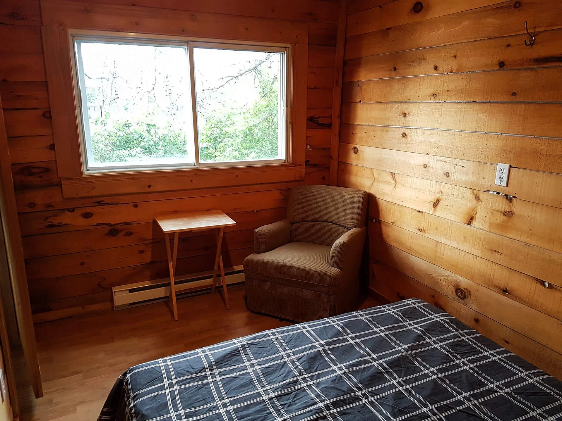 A bedroom with a bed, an armchair, and a small table next to a window overlooking the woods.