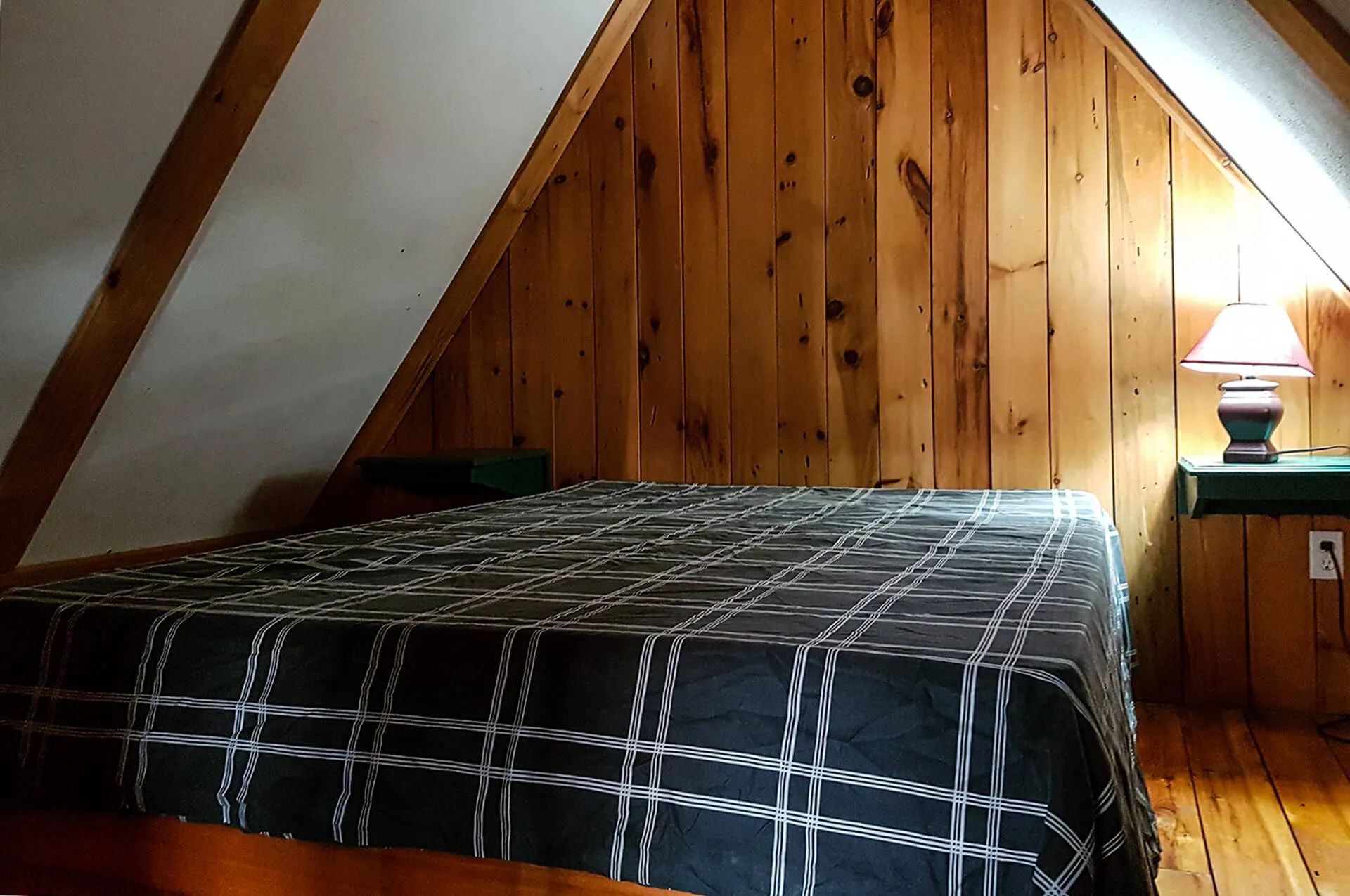 A bed under the slanted roof with a bedside lamp in the cabin.