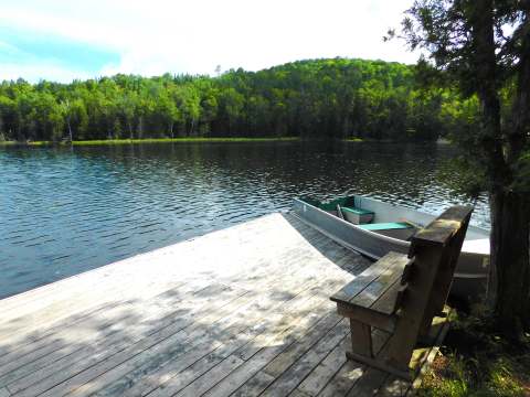 A boat is moored to the dock in front of the cabin with a view of the lake and the forest.