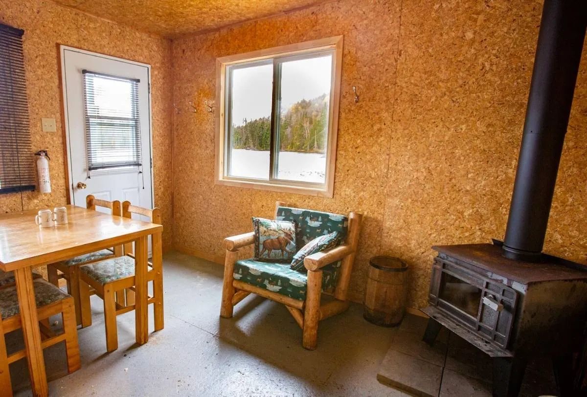 The interior of the cabin features a wooden table, a rustic armchair, and a wood-burning stove.