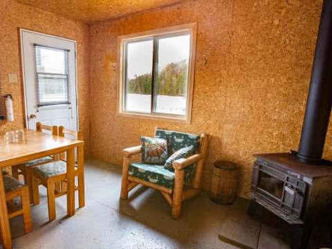 The interior of the cabin features a wooden table, a rustic armchair, and a wood-burning stove.