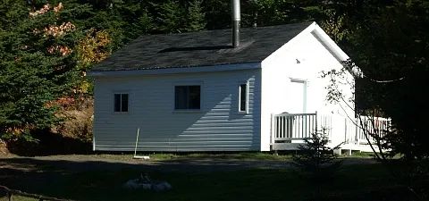 A white cottage with a porch and a chimney, located at the edge of the forest.