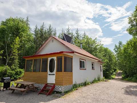 Vue extérieure du chalet avec sa véranda grillagée, sa table de pique-nique et son barbecue.