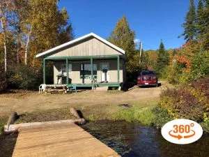 View of the Le Vison cabin with its covered porch, located by the water with a wooden dock.