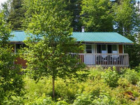 Le chalet L'Épervier avec son toit vert et sa galerie en bois est entouré par la forêt.