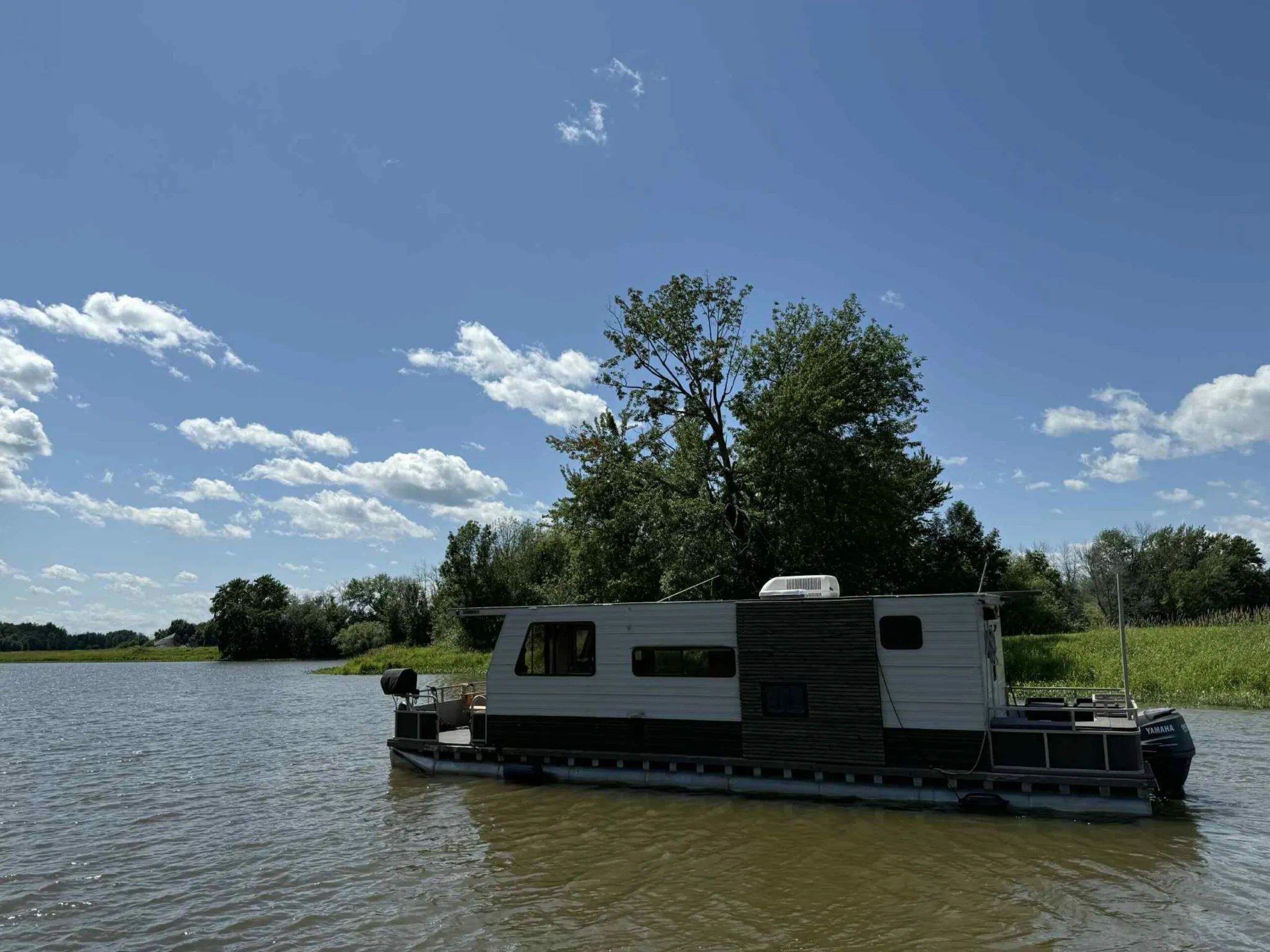 Le bateau-maison L'Eau-Tel flotte sur le plan d'eau entouré de verdure.