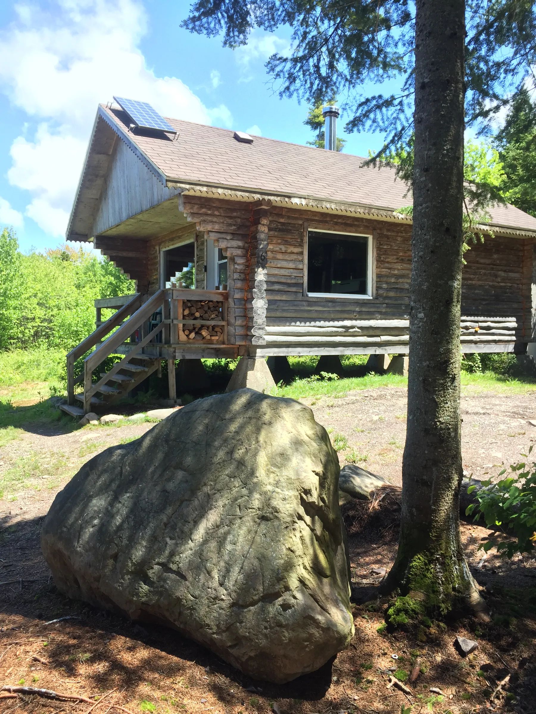 Vue latérale du chalet en bois avec ses escaliers et un grand rocher à proximité.