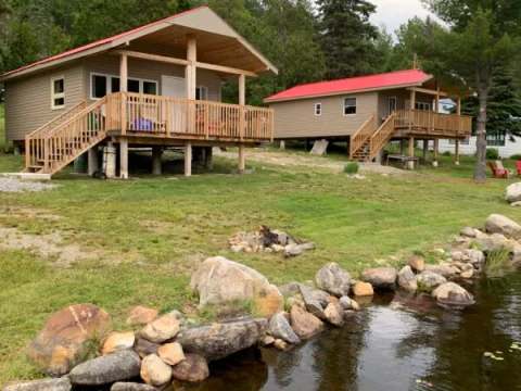 Two cabins with decks are located by the water in a wooded environment.