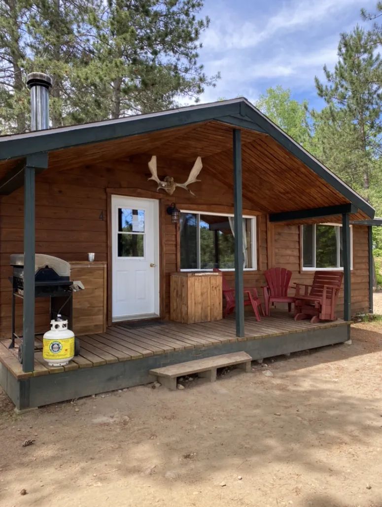 A view of the cabin front with its porch equipped with chairs, a barbecue, and natural wood finish.