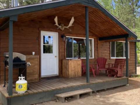 A view of the cabin front with its porch equipped with chairs, a barbecue, and natural wood finish.