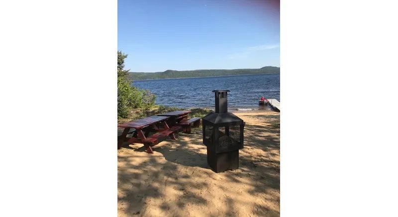 A picnic table and an outdoor fireplace are set up on the beach in front of the cabin.