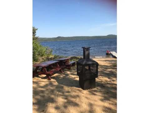 A picnic table and an outdoor fireplace are set up on the beach in front of the cabin.