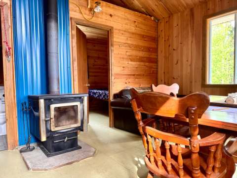 L'aire de séjour du chalet comprend un poêle à bois, une table en bois et un accès vers la chambre.