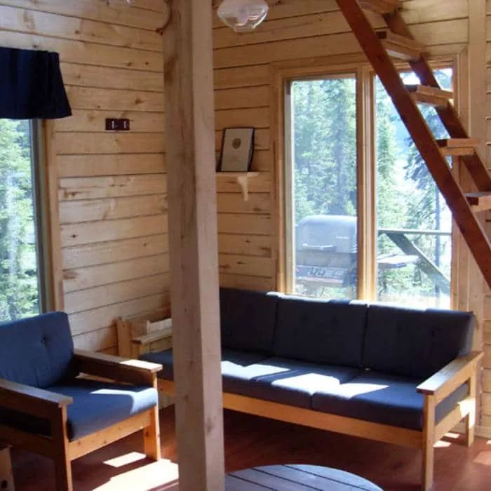 The cottage living room features a wooden sofa and armchair near a large window.