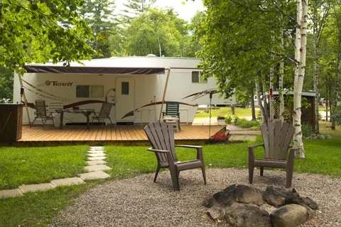 Emplacement de camping VR avec terrasse en bois, espace foyer extérieur et arbres matures.