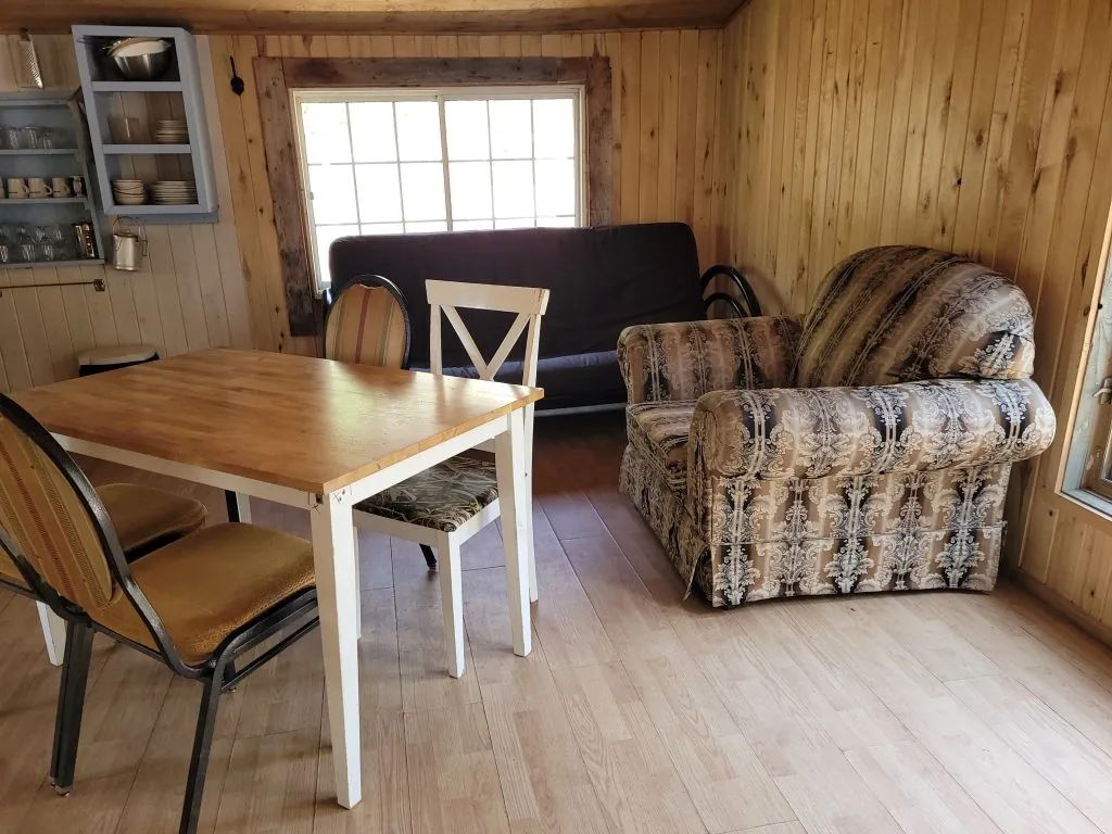 Coin repas et salon aménagé avec une table, des chaises et un fauteuil dans le Camp Malfait.