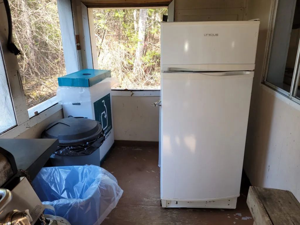 Un réfrigérateur et des bacs de recyclage sont installés sur la galerie fermée du chalet.