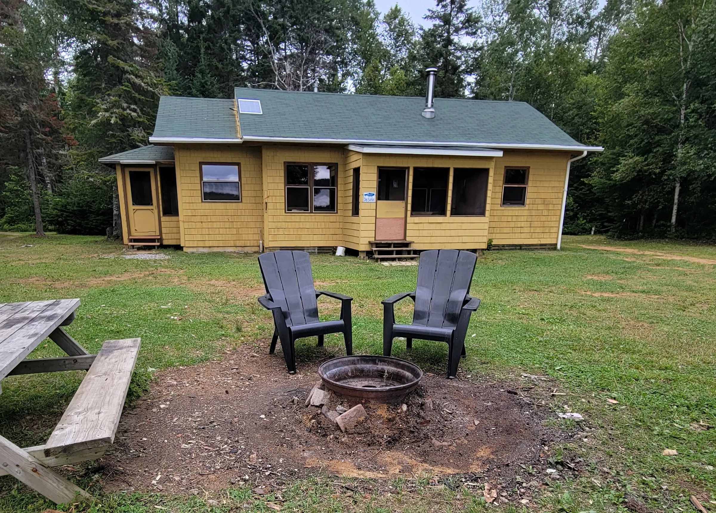 Vue du chalet Des Sables avec son foyer extérieur et ses chaises de détente sur le terrain.